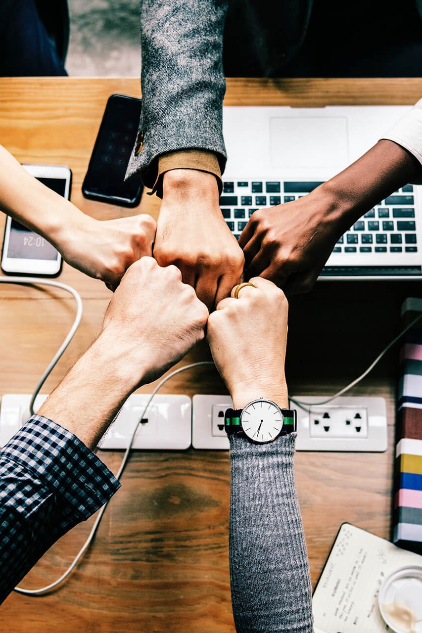 Fünf Personen stoßen über einem Schreibtisch mit Laptops und Smartphones die Fäuste zusammen – Symbol für Teamwork, digitale Kommunikation und Zusammenarbeit in der Krisenkommunikation.